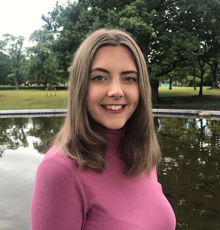 a smiling counsellor in a pink jumper stands in front of a pond in a park
